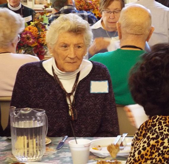 An elderly woman sits at a dining table in the crowded Parish Hall on All Saints Day, wearing a dark purple sweater.