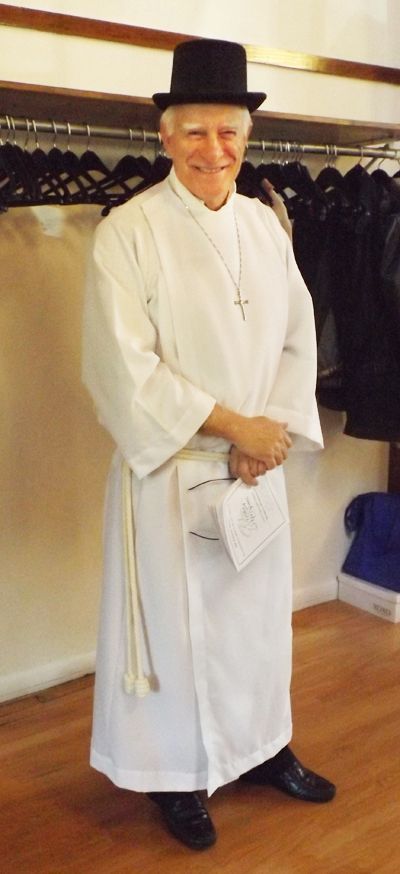 A chuckling male liturgical assistant in a white robe and black top hat stands in the Parish Hall with a coat rack behind him