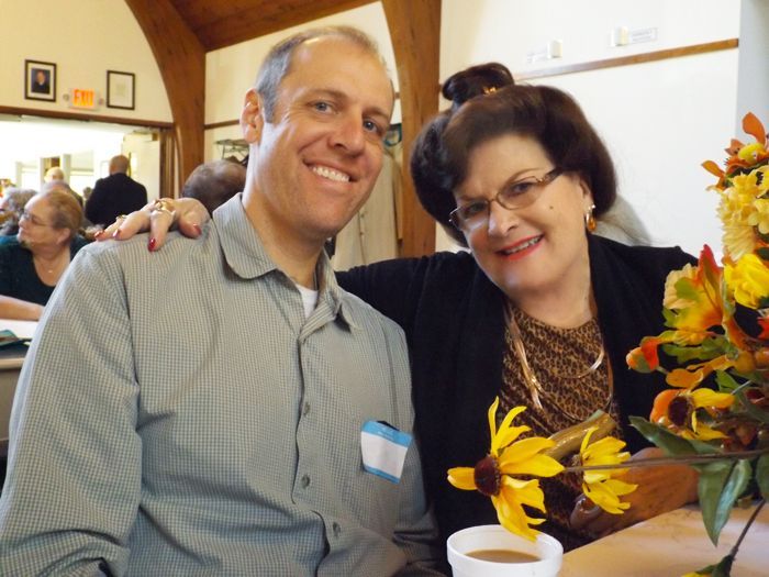 Two people smiling together at a table with yellow and orange flowers in the Parish Hall during Coffee Hour after service.