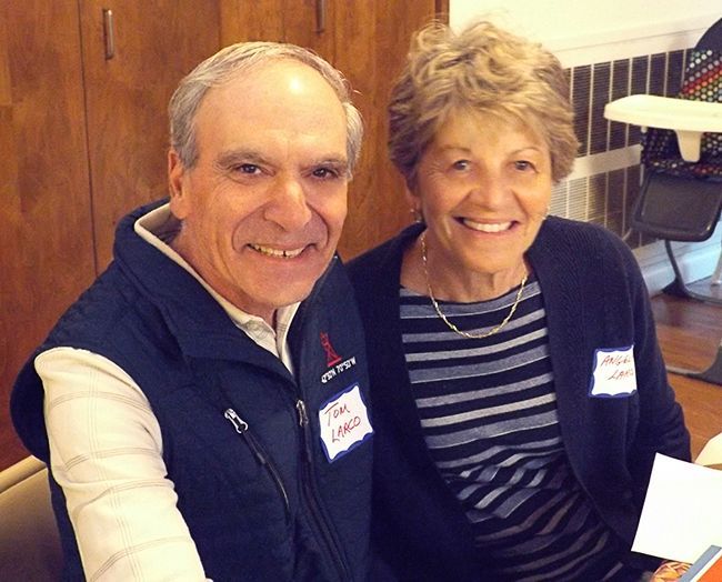 A man & a woman sit indoors, wearing name tags, in the warmly lit Parish Hall.