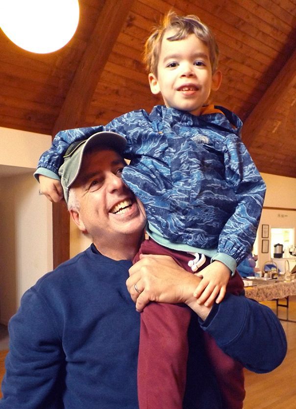 A man in a blue sweatshirt and baseball cap smiles while holding a young boy in a blue jacket on his shoulder in Parish Hall.