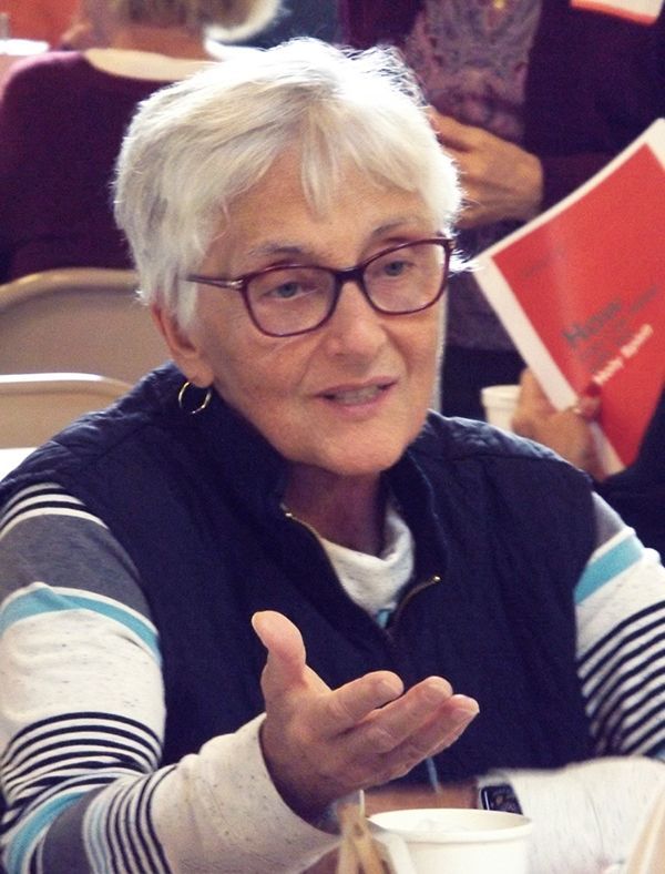 A woman with short white hair & glasses sits & speaks at a table, gesturing with one hand.