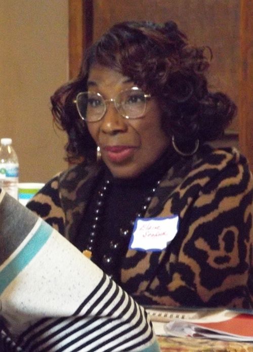 A black woman with curly hair & glasses sits at a table indoors, wearing a leopard-print sweater & a dark pearl necklace.