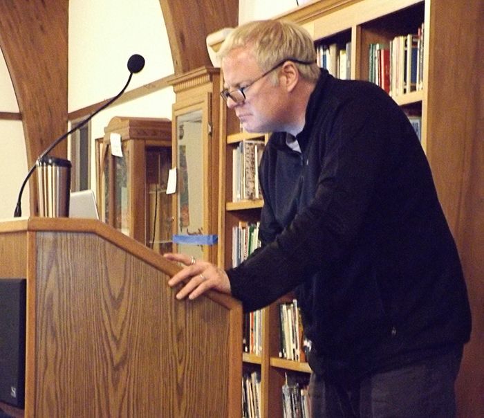 Pastor Charlie standing at a wooden podium in the Parish Hall with bookshelves behind him, fiddles on a laptop computer.