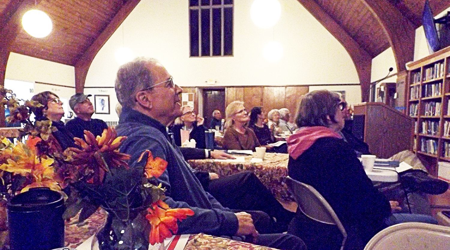 A large group of people sits listening to a speaker at tables in the Parish Hall, with a floral centerpiece and bookshelves