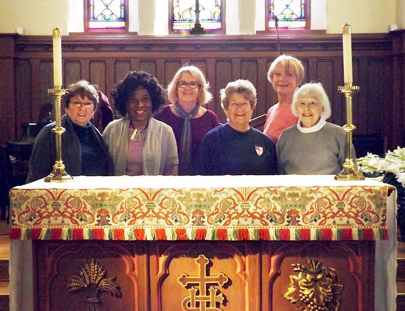 Six women from the church Altar Guild stand behind an ornate church altar, smiling, in a stained-glass sanctuary.