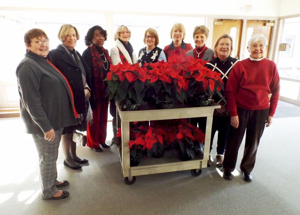 Nine women from our Altar Guild stand behind a cart of red poinsettias, in the bright church lobby.