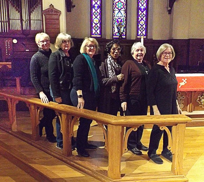 Six females on our church Altar Guild stand on a church chancel beside a wooden rail, with stained-glass windows behind them.