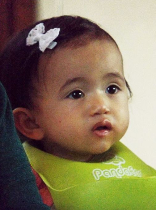 A baby in a green bib with a white bow in her hair, gazes ahead with a curious expression.