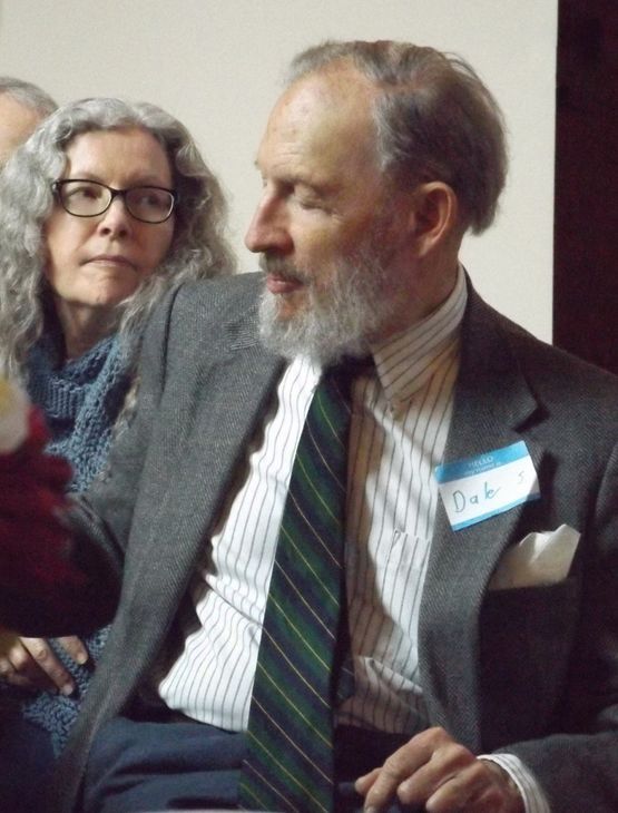 A bearded, grey-haired man in a dark suit w/a green striped tie sits near a woman w/long, wavy grey hair, at Annual Meeting.
