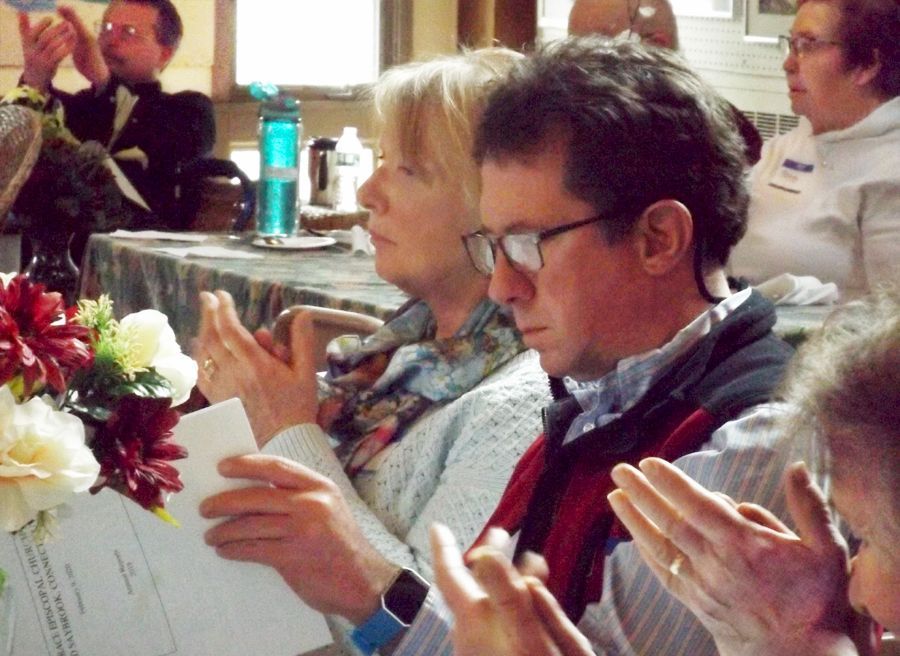 Groups of male & female parishioners sit at tables indoors, some holding programs & clapping, during a speech.