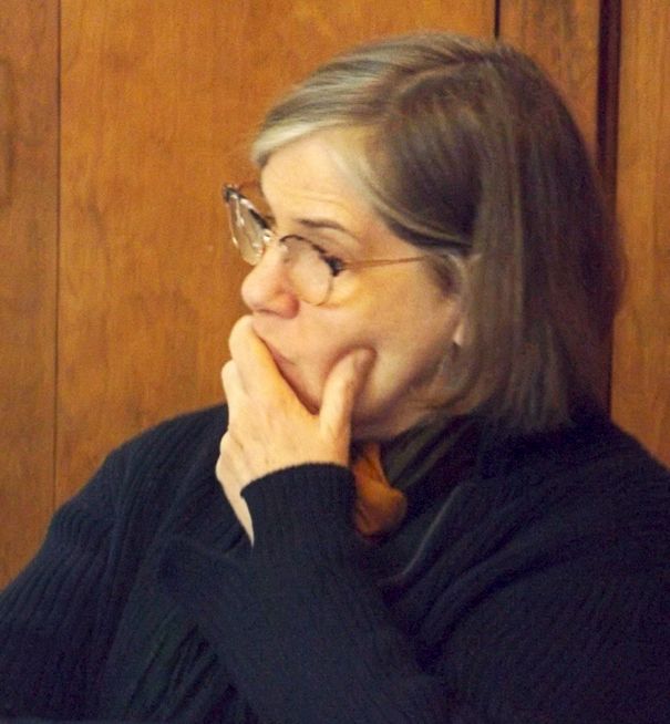 A woman wearing glasses in a wood-paneled room holds her hand to her face thoughtfully .