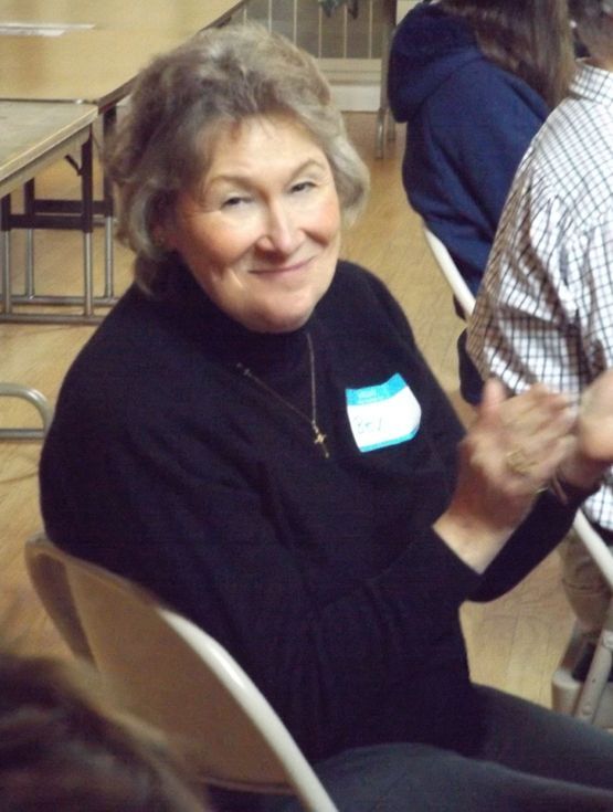 A smiling woman wearing a black turtleneck & a nametag, claps her hands while seated in a folding chair at the Annual Meeting