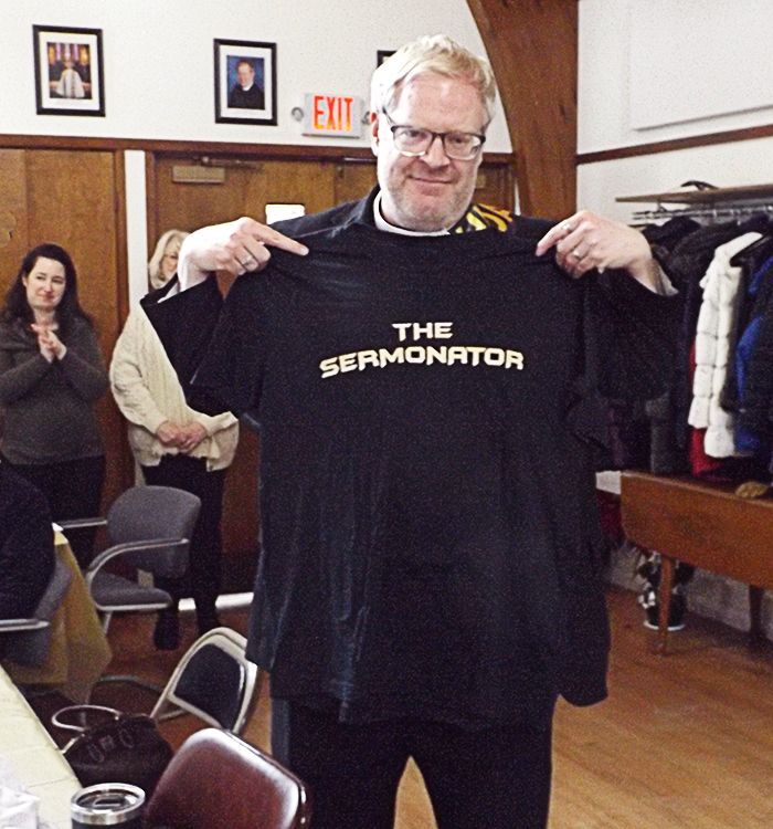 Pastor Charlie holds up a black T-shirt reading “THE SERMONATOR” in the Parish Hall with surrounding chairs and coats