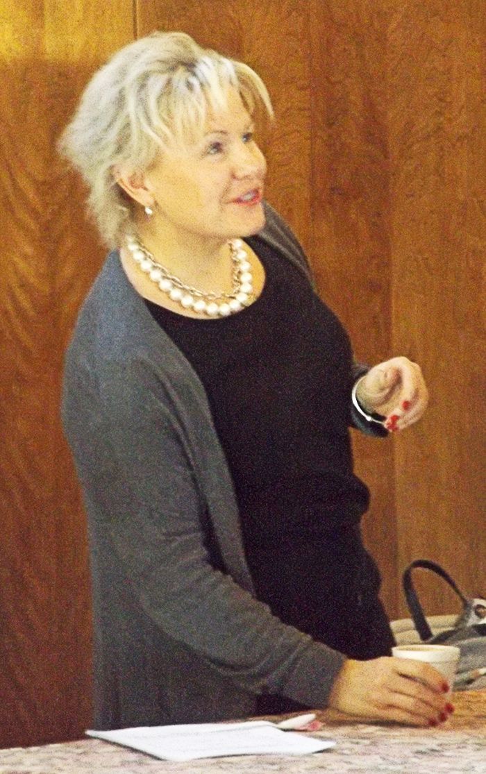 A woman wearing pearls, a black top & gray cardigan smiles & speaks to others while standing at a table.