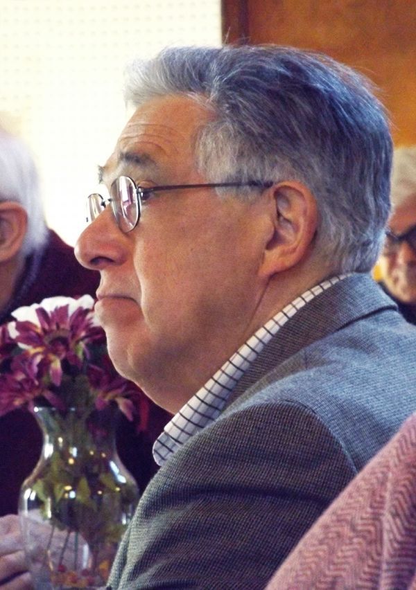 A grey-haired man w/glasses sits in profile wearing a gray suit-jacket, at a table w/purple flowers during the Annual Meeting