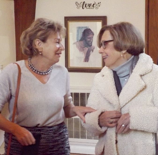 Two women smile & chat indoors, in front of a framed image of Jesus on the back wall.