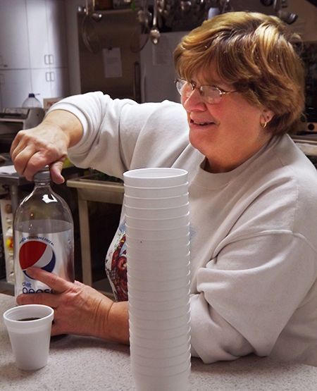 A smiling woman pours soda into a styrofoam cup, beside a stack of additional cups on a counter.