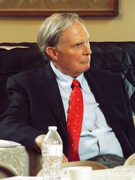 A well-dressed man in a suit & red tie sits slightly turned w/a serious expression while listening to a speaker