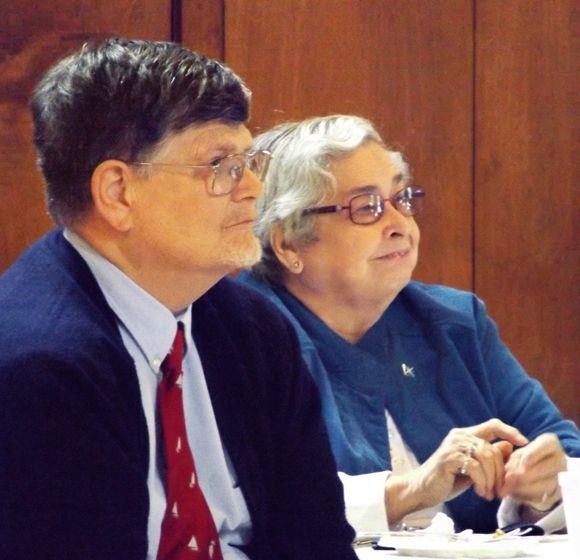 A husband & wife in glasses sit together
 at a conference table, listening attentively