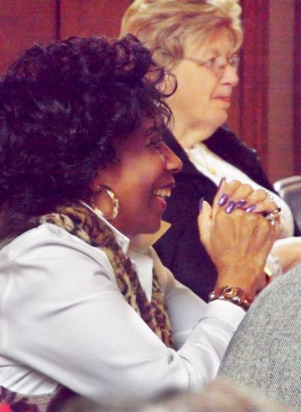 A black woman w/manicured purple nails & hoop-earrings laughs, while sitting w/her blond friend at the church Annual Meeting