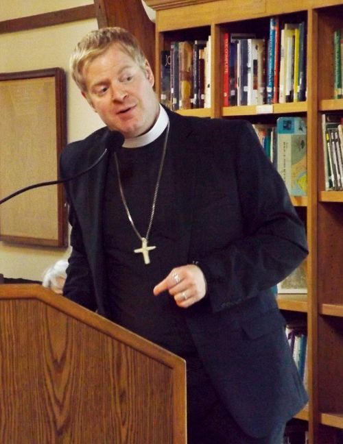 Pastor Charlie stands in clerical collar speaking at a podium, with bookshelves behind him.