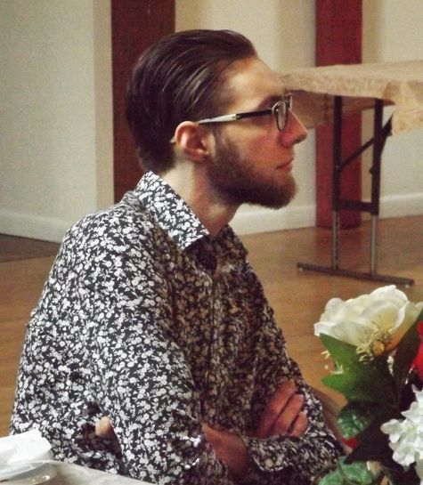 A bearded man in glasses & a patterned shirt sits indoors at a table w/flowers listening to a speaker at the Annual Meeting.