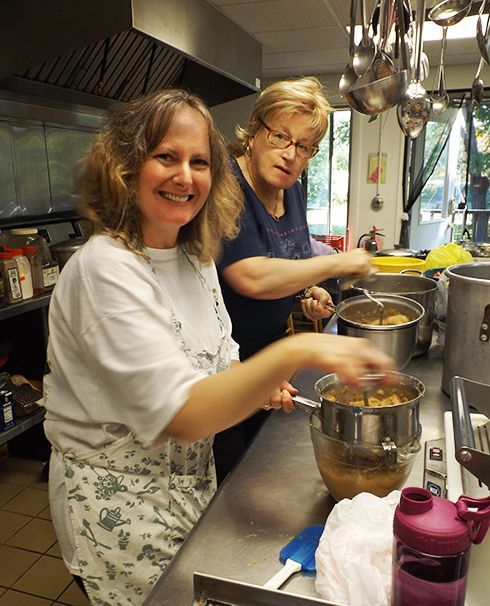 Two female parishioners cooking in the church's commercial kitchen, smiling while stirring pots at a stainless-steel counter