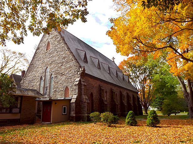 Our stone church with its steeply pitched roof stands in the grassy yard full of vibrant yellow and orange autumn foliage.