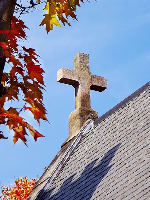A stone cross stands on a church roof against a blue sky, framed by autumn leaves.