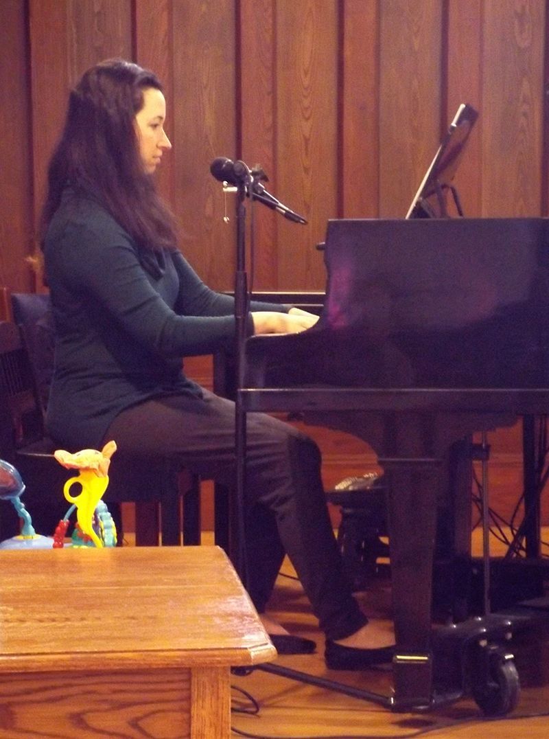 Our dark-haired female Music Minister sits paying piano & singing into a microphone, against the wood-paneled altar.