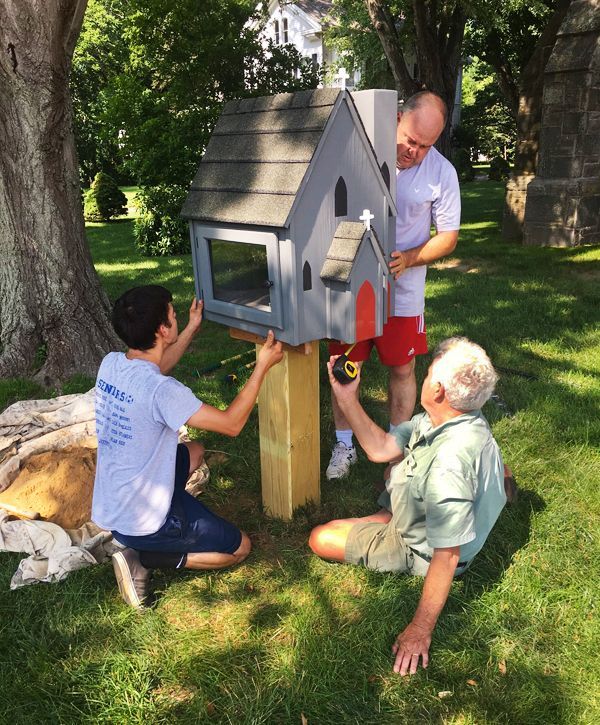 Three males install a gray little free library Bible Book Box in the sunny grassy churchyard.