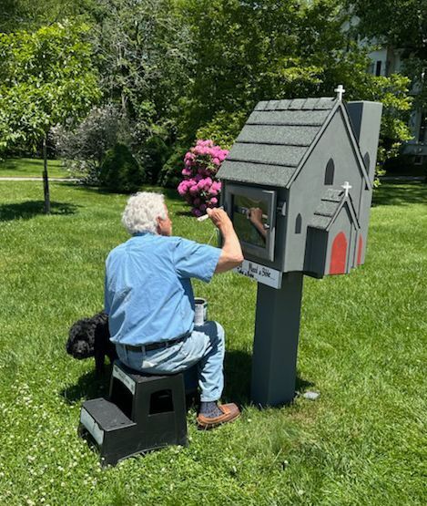 A man works on a little gray Bible Book Box in the sunny green churchyard with his black dog peeking out beside him.