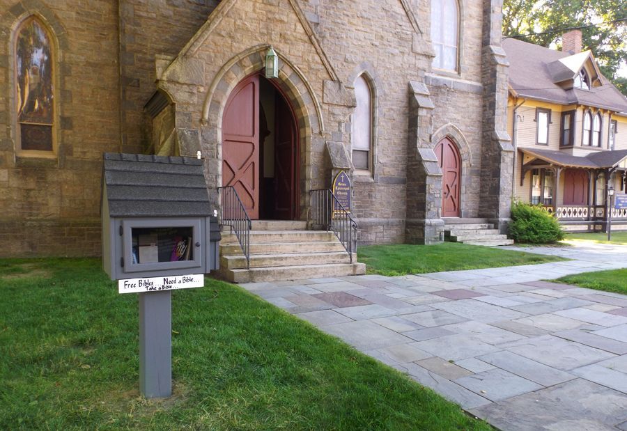 Stone church entrance with open arched red door, small gray Bible Book Box, and paved path on the lawn, with rectory in back