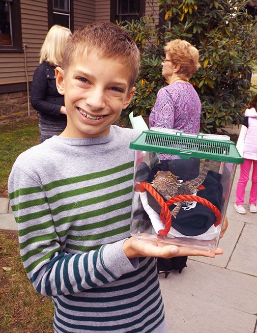 A smiling boy in a striped sweater holds a clear plastic pet carrier with a small bearded dragon inside