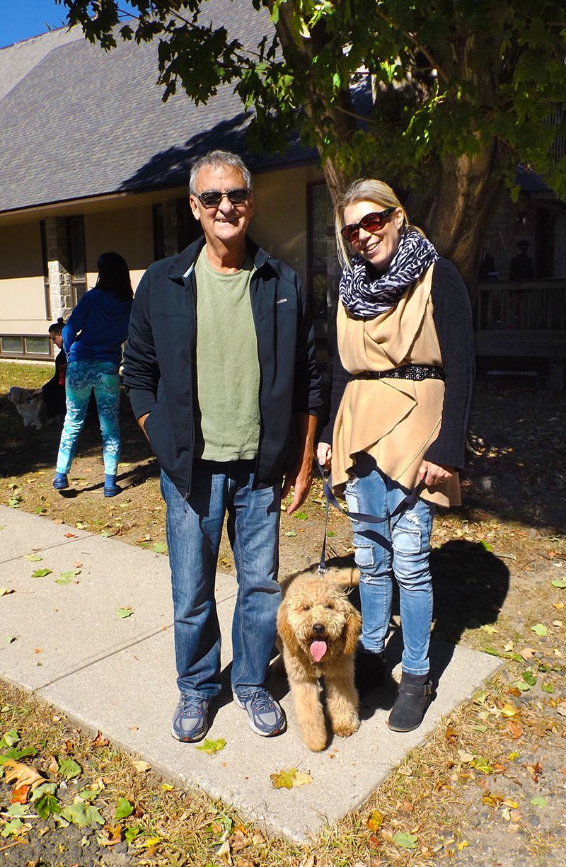 A couple in sunglasses stands outdoors in front of the church building w/their leashed dog