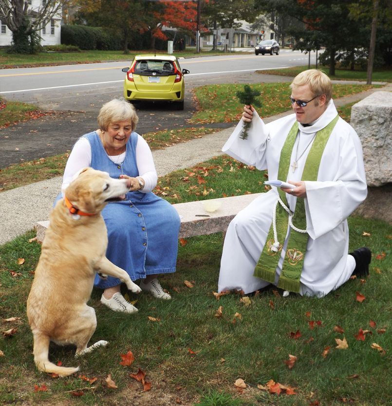 Pastor Charlie blesses a golden retriever beside a seated woman on the stone bench w/the street running behind her