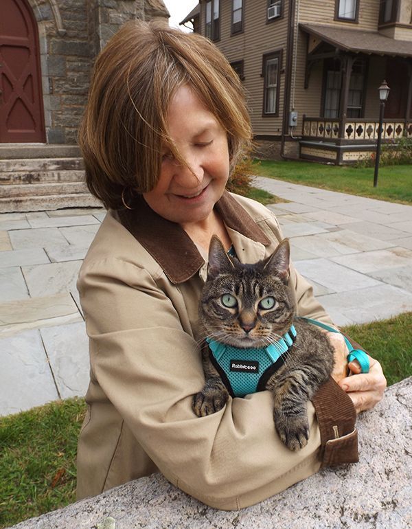 A woman in a tan jacket holds a brown tabby cat wearing a bright aqua harness outdoors in front of the stone church building.