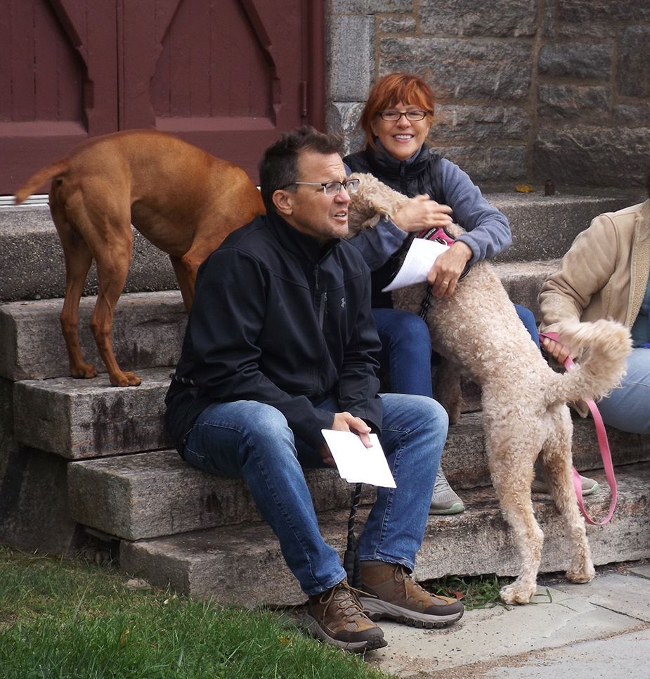 A man & a red-haired woman sit on the stone front steps of the church, waiting for an outdoor ceremony, w/2 large dogs