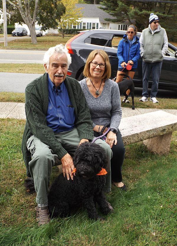 Older couple seated on a lawn bench with a black dog; people and a parked car in the background.