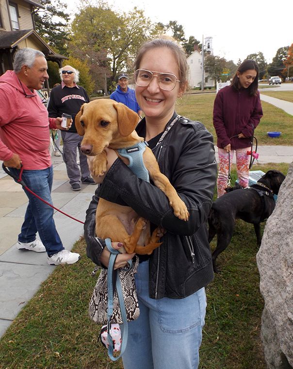 A smiling young woman in glasses holds her harnessed brown dog outdoors, w/several people & other dogs nearby.