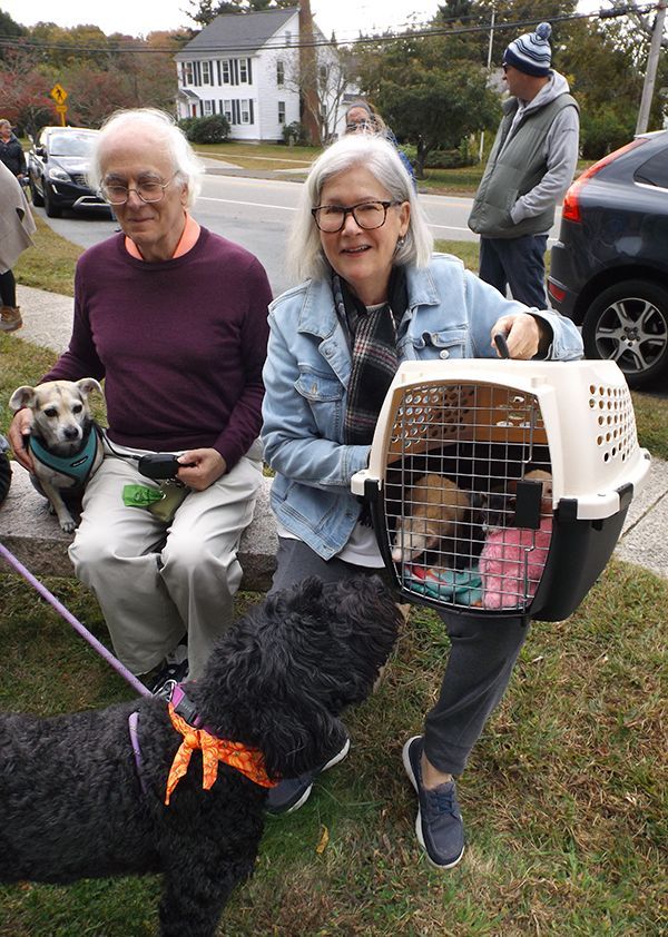 A husband & wife sit together w/their small, harnessed dog on a stone bench outside the church, holding a crate w/2 ferrets