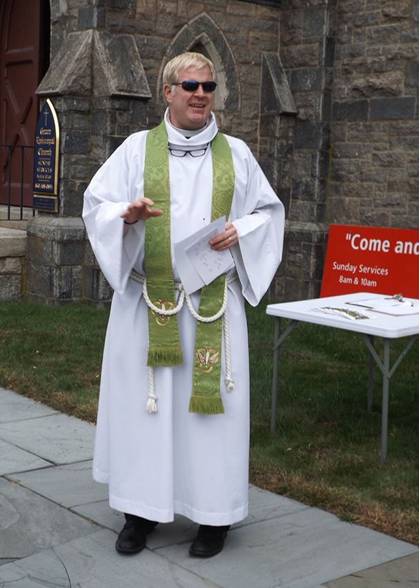 Pastor Charlie stands in white robes w/a green stole, on the sidewalk outside the stone church, holding papers & smiling