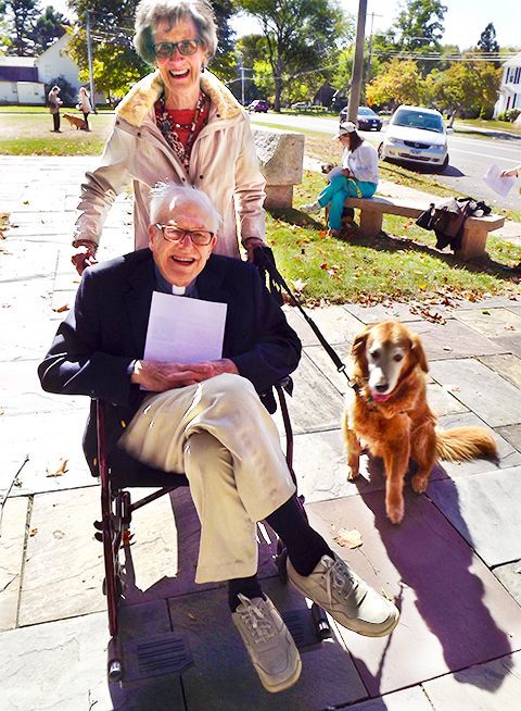 Elderly man in a wheelchair sits w/his smiling wife behind him & a dog beside them on a sunny sidewalk