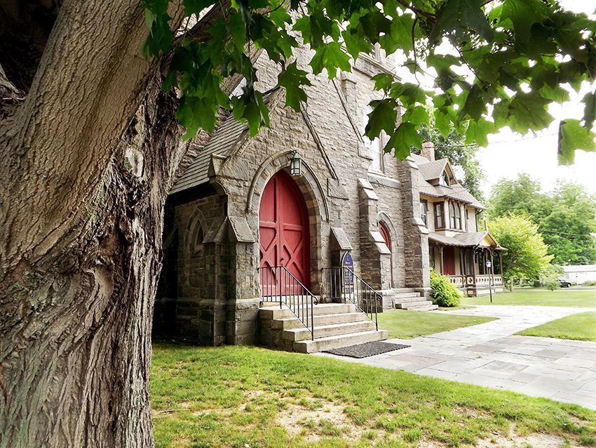 Stone church front-entrance with red doors framed by a leafy green tree on the lawn, beside a sidewalk with a rectory in back