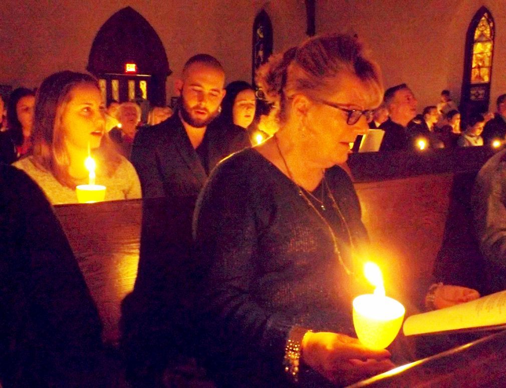 Parishioners in a dimly lit church hold glowing candles while singing during an evening candlelight service.