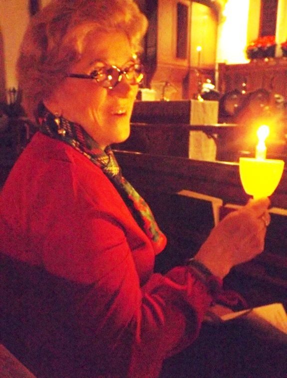 A woman wearing a red sweater sits in a dim church pew holding a lit candle while looking over her shoulder smiling