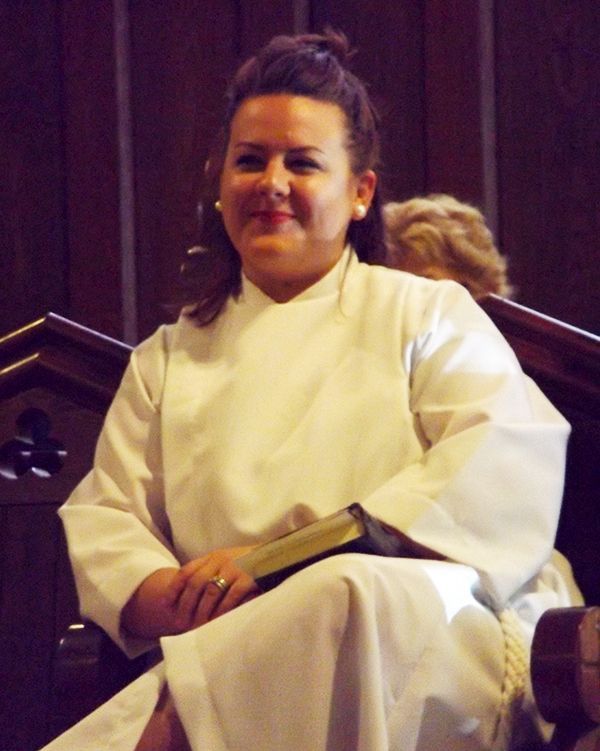 Female Eucharistic Minister in a white robe seated in the church sanctuary, smiling.