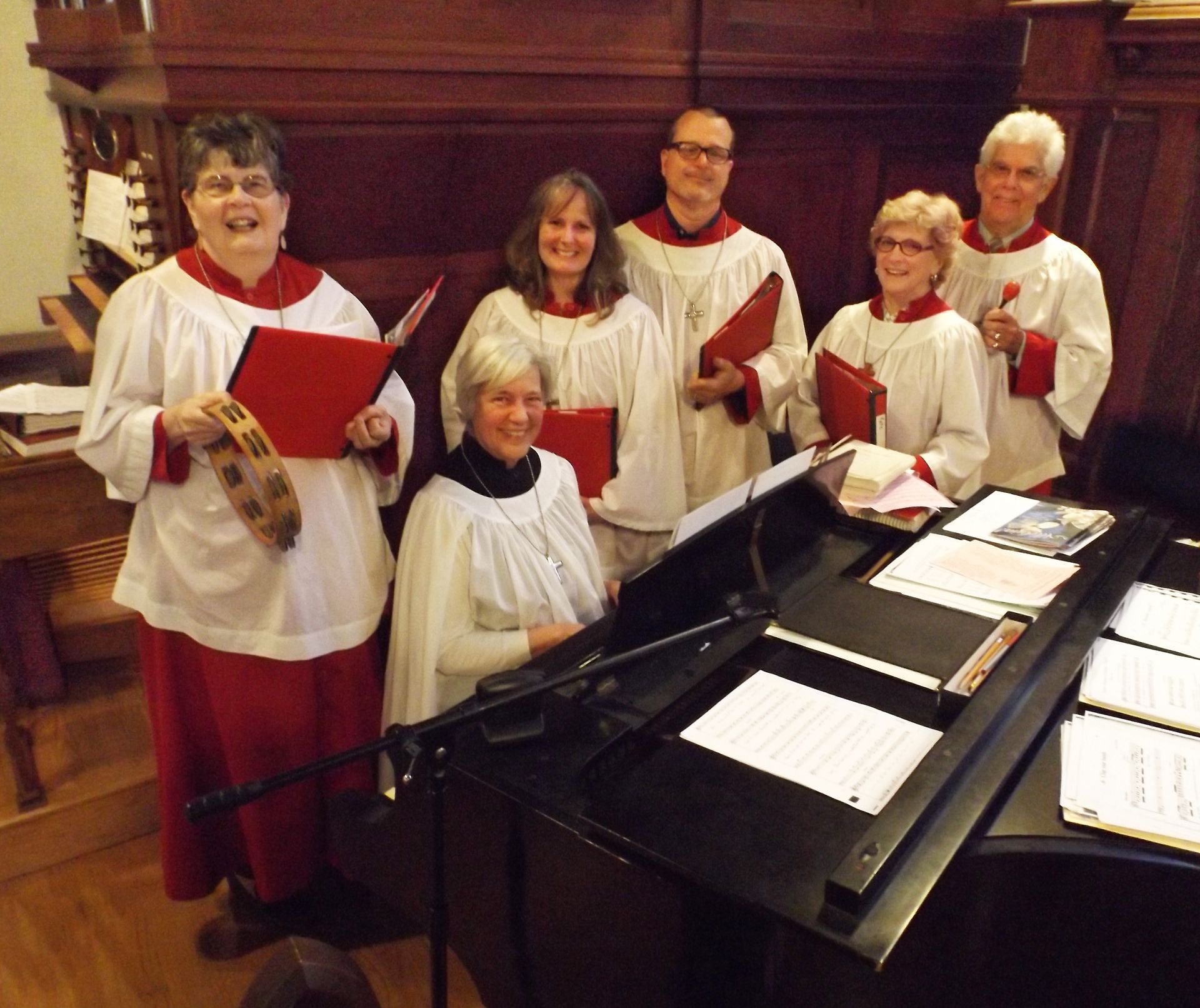 The church choir clusters around a piano together, in red & white robes, holding red music folders on the wooden altar