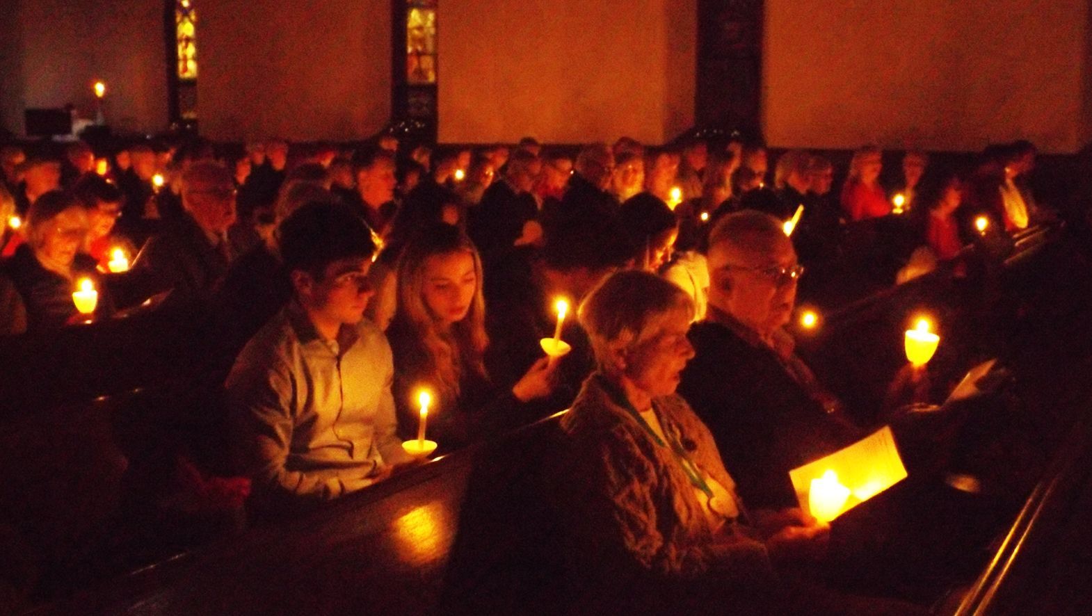The congregation holds glowing candles in the dark, during an indoor evening Christmas Eve candlelight service.
